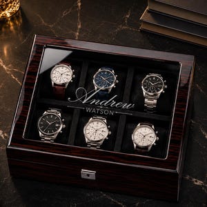 May include: A dark wood watch display case with a glass lid, containing six wristwatches. The case is engraved with the name "Andrew Watson". The watches feature different designs, with silver, black, and brown leather straps.