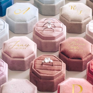 May include: A selection of velvet ring boxes in shades of pink, blue, and grey. Some boxes have gold lettering with initials, names, and dates. Several boxes display engagement rings with diamond detailing.