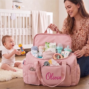 A woman is kneeling on the floor, holding a pink diaper bag filled with baby supplies, while a baby sits nearby, smiling and playing with a toy.