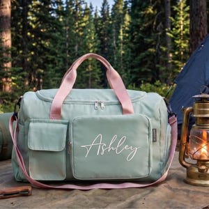 A green duffel bag with the name "Ashley" printed on it, along with camping gear such as a lantern and a sleeping bag, are displayed in a forest setting.