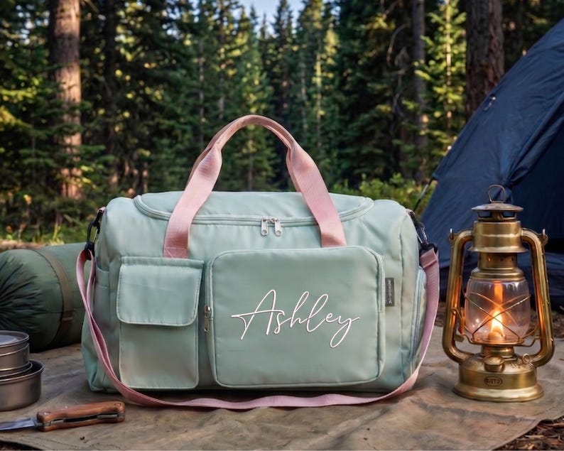 A green duffel bag with the name "Ashley" printed on it, along with camping gear such as a lantern and a sleeping bag, are displayed in a forest setting.