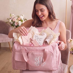 A woman in a pink dress is holding a pink bag with a personalized monogram, smiling as she looks inside the bag.