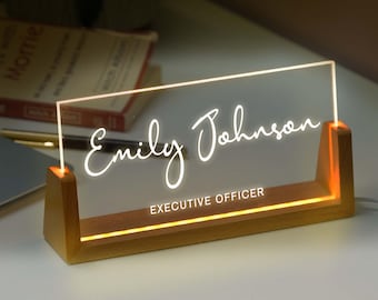 May include: A rectangular clear acrylic desk sign with a light-up base made of light-toned wood. The sign features the white illuminated text Emily Johnson in a script font and the text EXECUTIVE OFFICER in a sans-serif font.