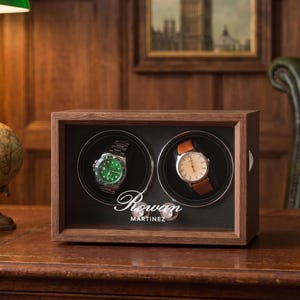 May include: A rectangular wooden watch winder box with a glass front, featuring two circular rotating compartments. Inside, a silver watch with a green face and a watch with a white face and brown leather strap are displayed. The text Rowan Martinez is printed in white.