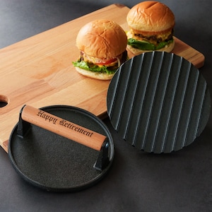 May include: Two round cast iron burger presses with wooden handles. One press features the engraved text Happy Retirement on the handle. The other press shows a ribbed underside. The items are placed near two cheeseburgers on a wooden board.