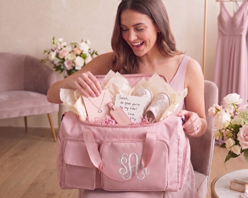 A woman in a pink dress is holding a pink bag with a personalized monogram, smiling as she looks inside the bag.