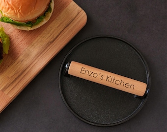 May include: A round black cast iron burger press with a light-colored wooden handle. The handle is engraved with the text "Enzo's Kitchen" in a dark font. The press features a flat circular base and a horizontal handle attached to the top.