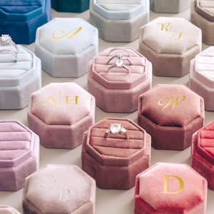May include: A collection of velvet ring boxes in shades of blue, pink, and red. Each octagonal box displays a gold initial or a ring. The boxes are arranged in a grid, highlighting the range of colours and designs available.
