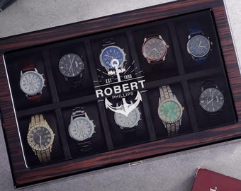 May include: A rectangular wooden watch display case with a dark wood grain finish and a glass lid. The lid features a white graphic of an anchor and the text "EST 1990 ROBERT PHILLIPS". Inside are ten individual compartments, each containing a wristwatch.