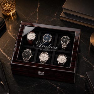 May include: A rectangular dark wood watch box with a glass lid featuring the engraved name Andrew Watson. The interior contains six individual compartments, each holding a different wristwatch with various dial colors and metal or leather bands.