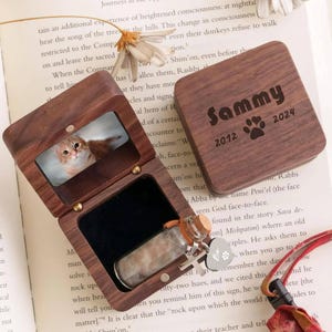 May include: A wooden keepsake box with a photo of a cat inside, and a separate lid engraved with the name "Sammy", paw prints, and the years 2012 and 2024. A small glass vial with a cork stopper and a charm are inside.
