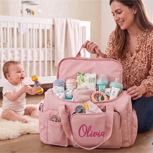 A woman is kneeling on the floor, holding a pink diaper bag filled with baby supplies, while a baby sits nearby, smiling and playing with a toy.