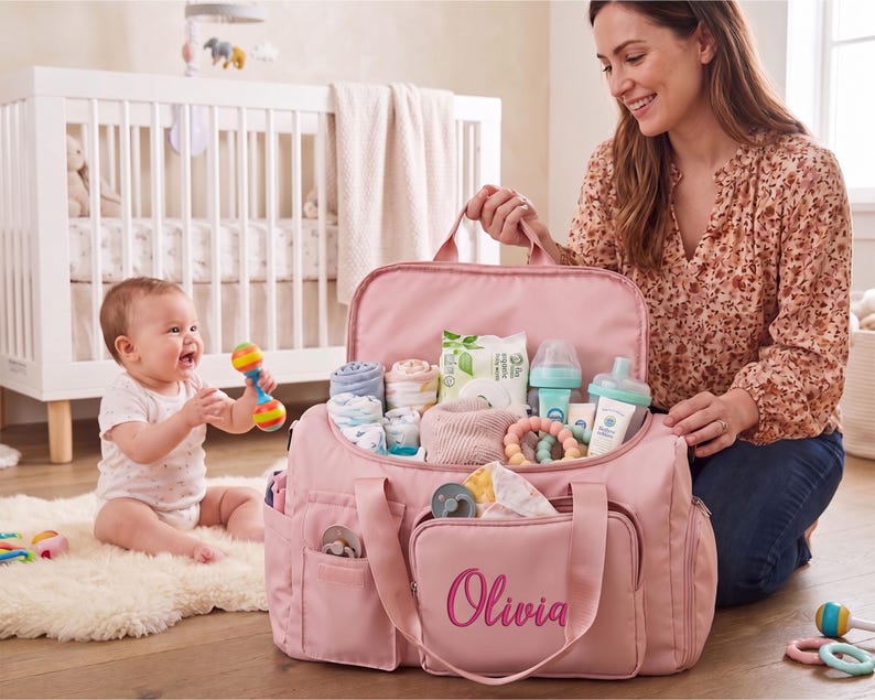 A woman is kneeling on the floor, holding a pink diaper bag filled with baby supplies, while a baby sits nearby, smiling and playing with a toy.