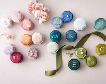 Könnte beinhalten: Eine Sammlung achteckiger Samt-Ringkästchen in verschiedenen Farben wie Rosa, Blau, Grün und Lila. Jede Schachtel ist mit Monogrammen, Namen oder Daten in Gold- oder Silberfolie versehen. Einige Schachteln sind geöffnet und zeigen Ringe.