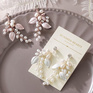 May include: Two pairs of floral-inspired drop earrings featuring white freshwater pearls, small clear crystals, and metallic leaves. One pair is set on rose gold wire, while the other is on silver wire, displayed on a card labeled HUNDRED HEARTS.