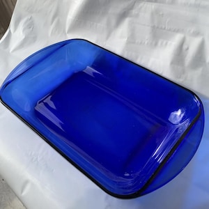 Puede incluir: Una fuente de horno rectangular de vidrio azul cobalto con esquinas redondeadas y asas integradas en ambos extremos. El recipiente presenta una superficie lisa y translúcida con un color azul intenso y vibrante en toda su estructura.