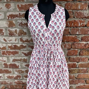 May include: White sleeveless dress with a floral pattern in red and blue. The dress has a V-neckline, a drawstring waist, and a knee-length skirt. The dress is displayed on a black mannequin.