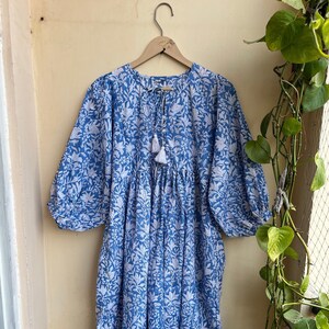 May include: A blue and white floral print dress with a gathered waist and 3/4 sleeves. The dress has a tie at the neckline with a tassel.