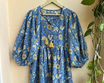 May include: A blue floral dress featuring yellow and white flowers. The dress has long, puffed sleeves, a round neckline, and a button-down front. Two yellow tassels adorn the neckline. The dress is suspended from a wooden hanger.