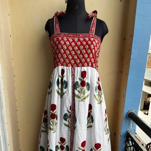 Könnte beinhalten: Ein weißes Baumwollkleid mit rotem Blumendruck. Das Kleid hat einen quadratischen Ausschnitt und dünne Träger, die an den Schultern gebunden werden. Der Rock ist voll und fließend.