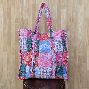 以下が含まれることがあります： ピンク、青、緑、黄色の花柄と幾何学模様が混ざったパッチワークのトートバッグ。バッグは長方形で、2つのハンドルが付いており、キルティング生地で作られています。端はピンクのボーダーでトリミングされています。
