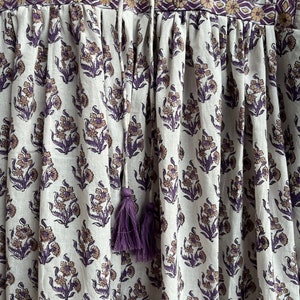 May include: A white cotton dress with a purple floral print and a purple tassel tie. The dress has a gathered skirt and a fitted bodice.