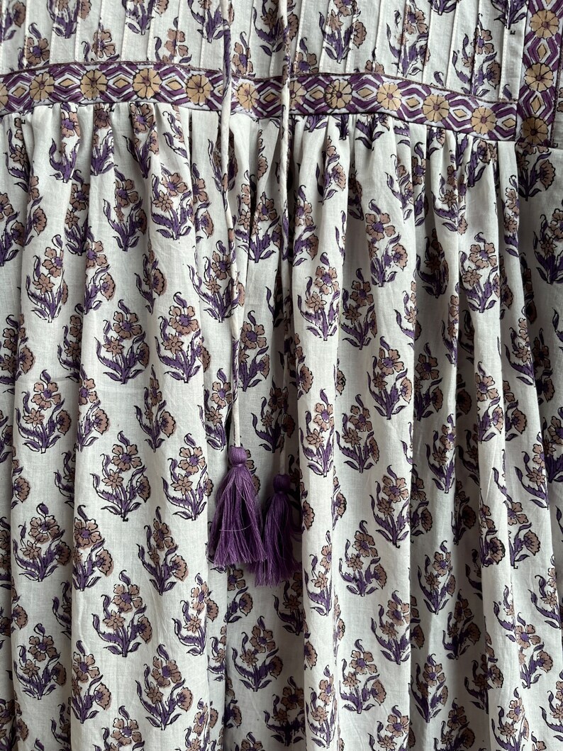 May include: A white cotton dress with a purple floral print and a purple tassel tie. The dress has a gathered skirt and a fitted bodice.