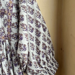 May include: A white cotton dress with a floral print in shades of purple and brown. The dress has a gathered bodice and long sleeves with elastic cuffs.