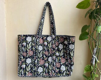Op de afbeelding: Een gewatteerde draagtas met een bloemenpatroon in wit, roze en groen op een marineblauwe achtergrond. De tas heeft bijpassende handvatten en een decoratieve rand. De tas hangt aan een beige muur.