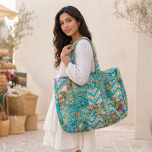 Könnte beinhalten: Eine große, gesteppte Tragetasche mit einem türkis-, blau- und floralen Muster. Die Tasche hat ein Zick-Zack-Design und wird von einer Person getragen, die ein weißes Kleid trägt. Die Tasche hat einen passenden Riemen.