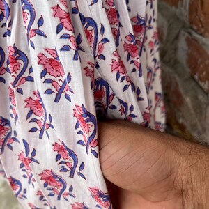 May include: Close-up of white fabric with a floral pattern in pink and blue. The design features stylized flowers and leaves. The fabric is draped, and a hand is visible in the pocket.