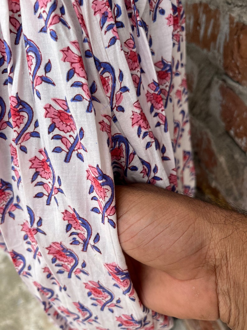 May include: Close-up of white fabric with a floral pattern in pink and blue. The design features stylized flowers and leaves. The fabric is draped, and a hand is visible in the pocket.