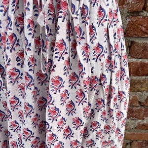 May include: White fabric with a repeating floral pattern in pink and blue. The fabric is gathered at the top with a drawstring. The background is a brick wall. The floral design is a stylized depiction of flowers and leaves.
