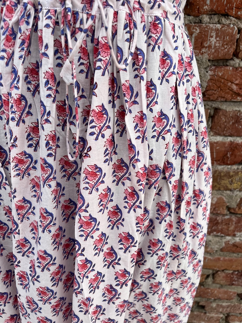 May include: White fabric with a repeating floral pattern in pink and blue. The fabric is gathered at the top with a drawstring. The background is a brick wall. The floral design is a stylized depiction of flowers and leaves.