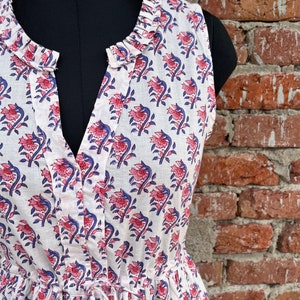 May include: Sleeveless white dress with a floral pattern in red and blue. The dress features a ruffled neckline, a V-neck, and a drawstring waist. The fabric is lightweight and appears to be cotton.