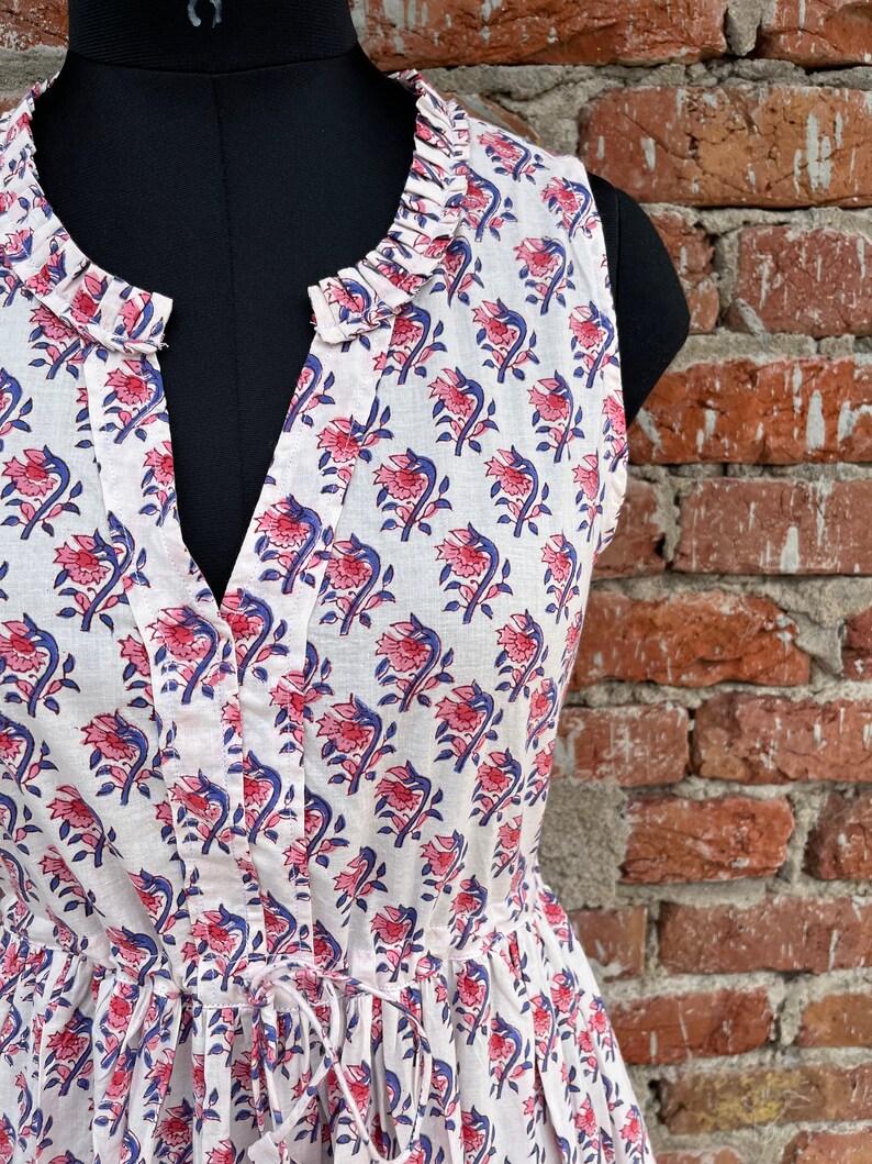 May include: Sleeveless white dress with a floral pattern in red and blue. The dress features a ruffled neckline, a V-neck, and a drawstring waist. The fabric is lightweight and appears to be cotton.