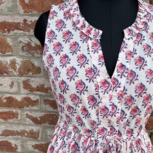 May include: White sleeveless dress with a floral pattern in pink and blue. The dress features a V-neckline, a drawstring waist, and a ruffled collar. The dress is displayed on a black mannequin against a brick wall.