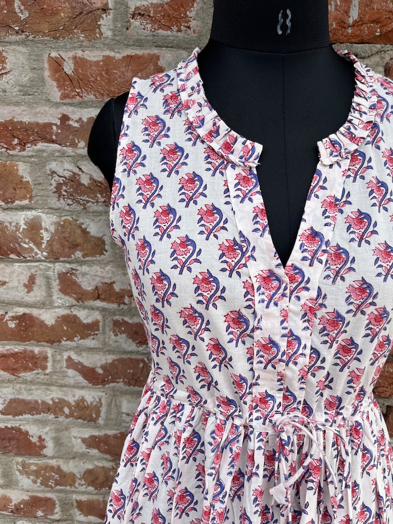May include: White sleeveless dress with a floral pattern in pink and blue. The dress features a V-neckline, a drawstring waist, and a ruffled collar. The dress is displayed on a black mannequin against a brick wall.