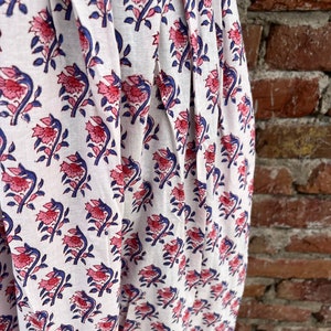 May include: White fabric with a repeating floral pattern in pink and blue. The flowers have detailed petals and are set against a white background. The fabric is draped, showing folds and texture, with a brick wall in the background.