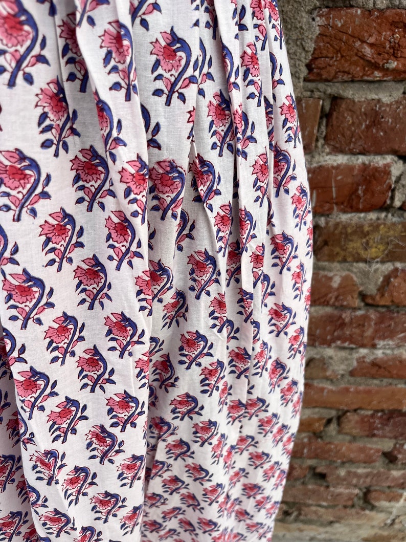 May include: White fabric with a repeating floral pattern in pink and blue. The flowers have detailed petals and are set against a white background. The fabric is draped, showing folds and texture, with a brick wall in the background.