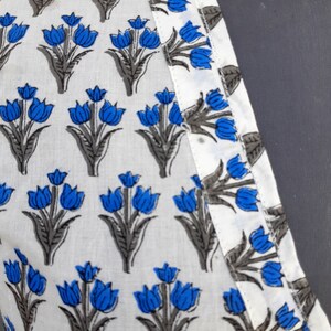 May include: White fabric with a blue and gray floral print. The fabric has a white seam along the edge.