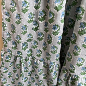 Op de afbeelding: Een grijze katoenen stof met een herhalend patroon van blauwe en groene bloemen. De stof heeft een gerimpelde rand.