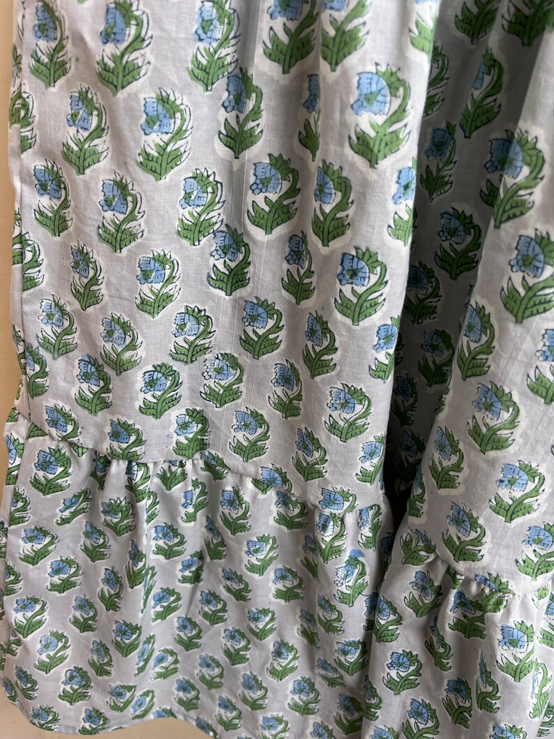 Op de afbeelding: Een grijze katoenen stof met een herhalend patroon van blauwe en groene bloemen. De stof heeft een gerimpelde rand.