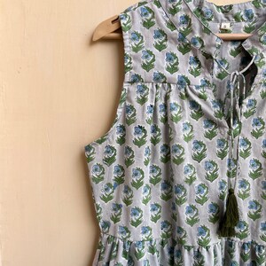 Op de afbeelding: Een mouwloze grijze jurk met een bloemenprint in tinten blauw en groen. De jurk heeft een strik aan de halslijn en een kwast aan de onderkant.