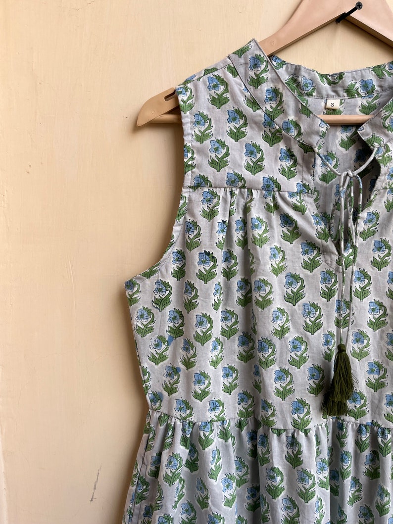 Op de afbeelding: Een mouwloze grijze jurk met een bloemenprint in tinten blauw en groen. De jurk heeft een strik aan de halslijn en een kwast aan de onderkant.
