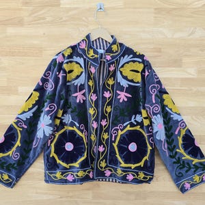 Può includere: Giacca in velluto blu scuro con ricami floreali intricati in giallo, rosa e azzurro. La giacca presenta un colletto rotondo e maniche lunghe, con una chiusura a bottoni. Il ricamo include grandi disegni circolari e delicati motivi floreali.