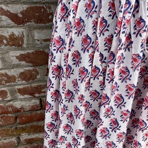 May include: White fabric with a repeating floral pattern in pink and blue. The flowers have a detailed design, and the blue elements create a contrast against the white background. The fabric is draped, suggesting it's part of a garment or textile.