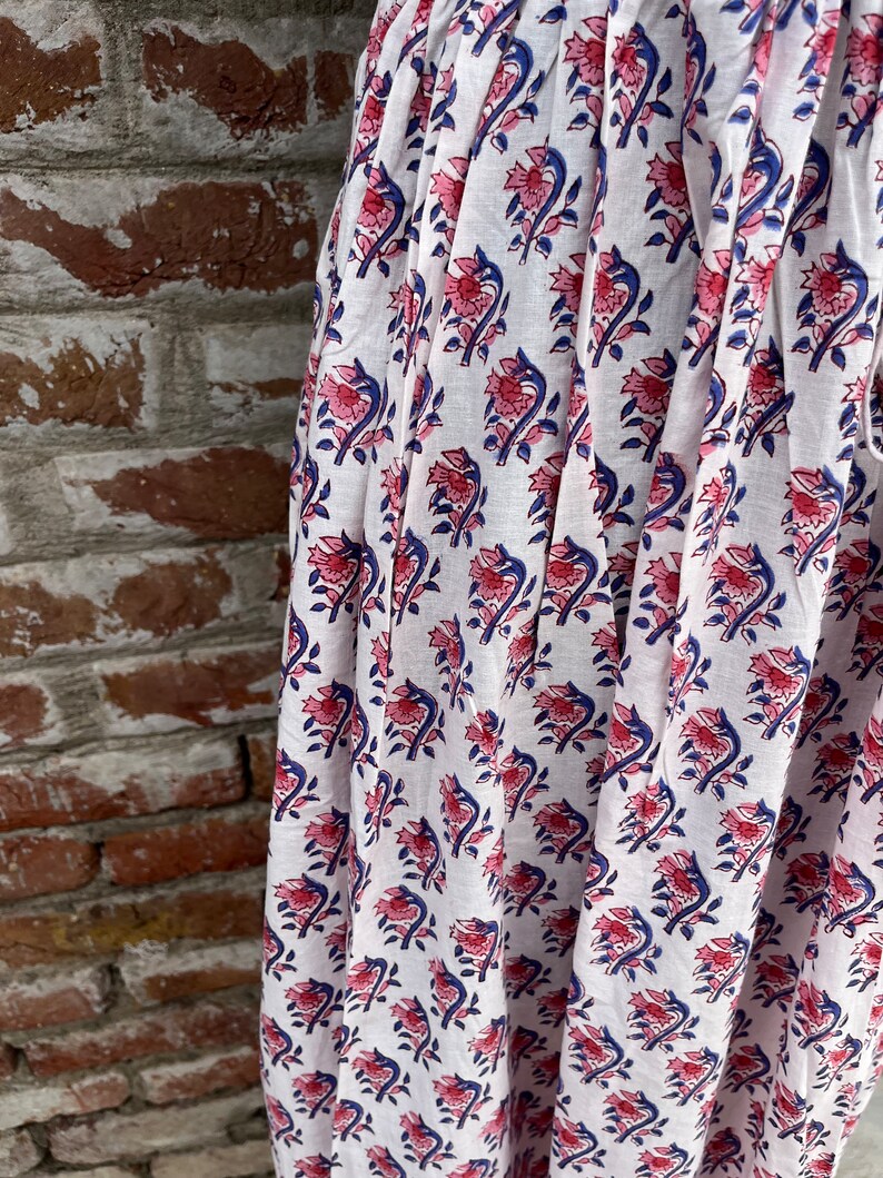 May include: White fabric with a repeating floral pattern in pink and blue. The flowers have a detailed design, and the blue elements create a contrast against the white background. The fabric is draped, suggesting it's part of a garment or textile.