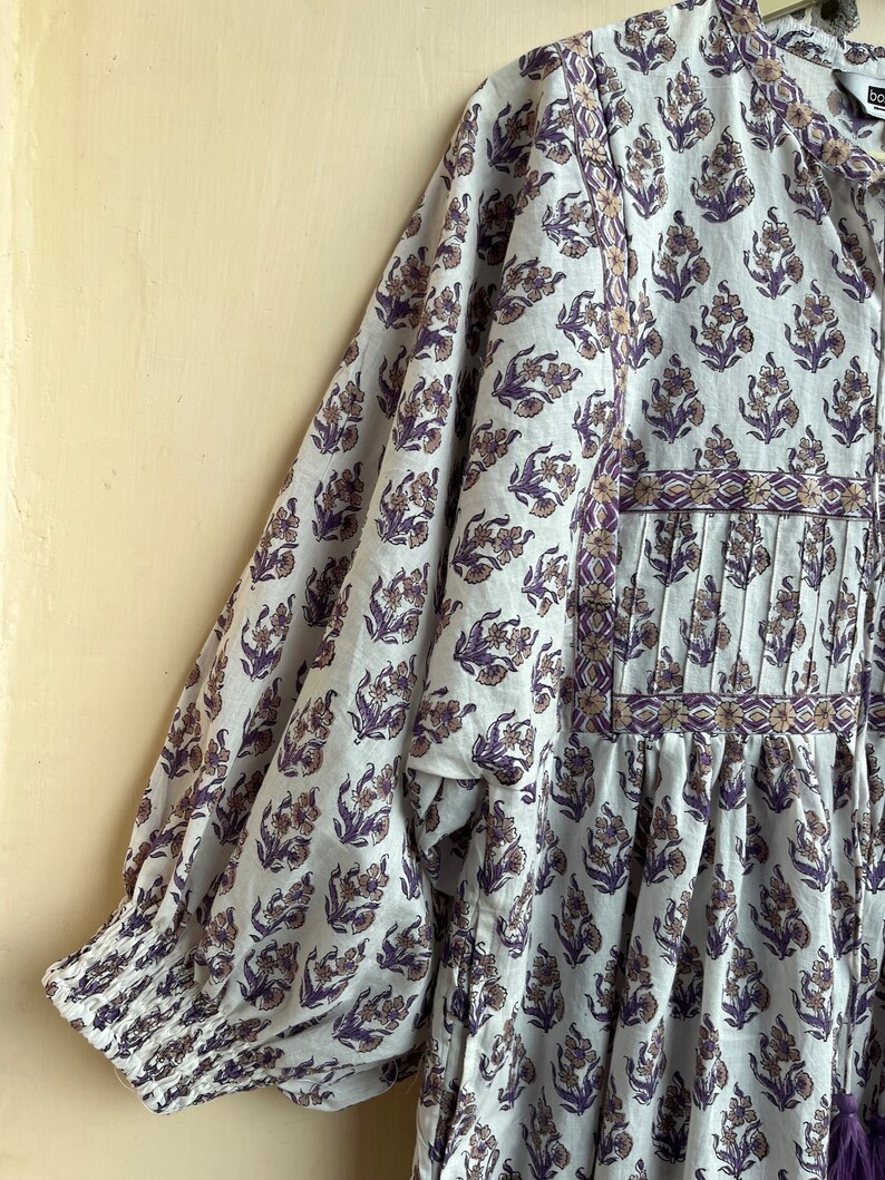 May include: A white cotton dress with a purple floral print and a gathered skirt. The dress has a round neckline and 3/4 sleeves with elastic cuffs.