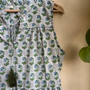 Op de afbeelding: Een grijs, mouwloos jurkje met een bloemenprint in blauwe en groene tinten. Het jurkje heeft een strik aan de halslijn en een kwast.